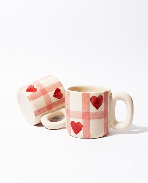 Two ceramic mugs with a pink and cream plaid pattern and red heart details, displayed on a clean white background.