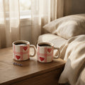 Two ceramic mugs with a pink and white gingham pattern and red hearts, filled with coffee and placed on a wooden bedside table next to a bed with beige linens.