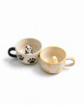 Two ceramic mugs on a white background. The left mug is cream with a black handle and black paw print, containing a small panda figurine.