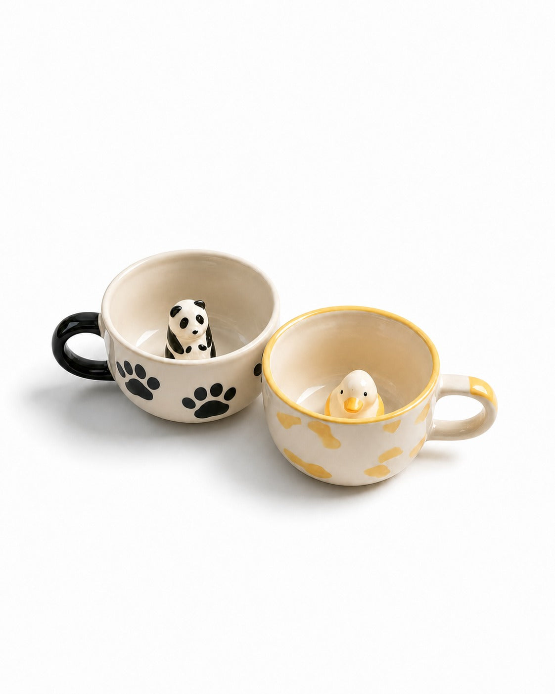 Two ceramic mugs on a white background. The left mug is cream with a black handle and black paw print, containing a small panda figurine.