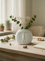 White ceramic vase with face design on a wooden surface with greenery