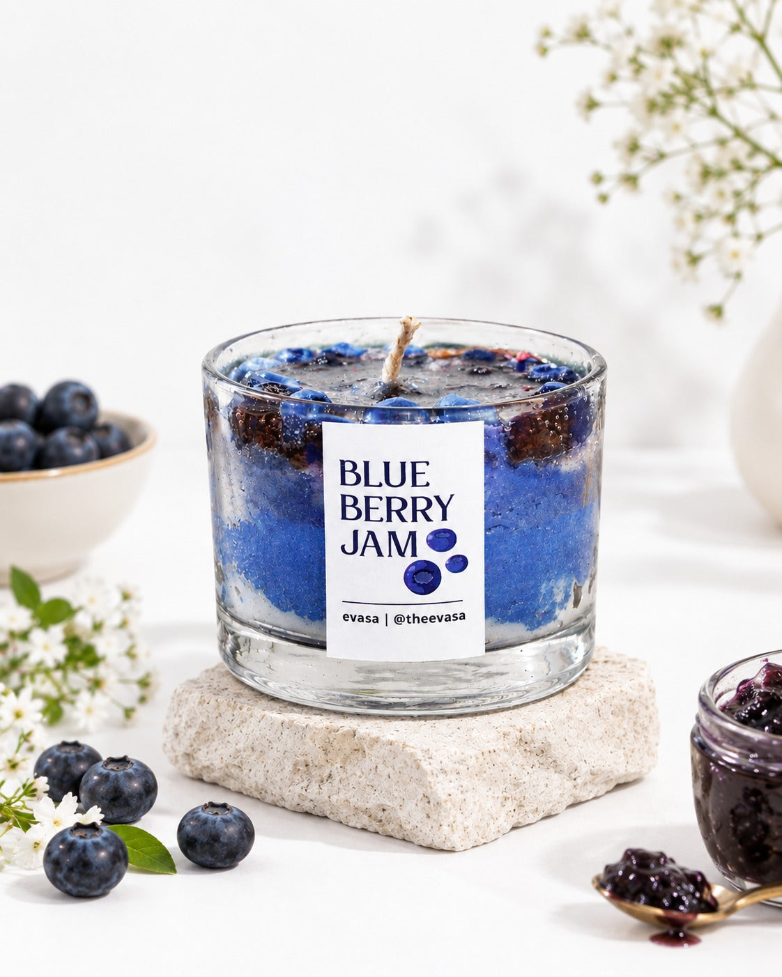 A glass jar candle with a wick and a white label reading 'BLUE BERRY JAM' sits on a stone coaster. The candle wax is deep blue with visible blueberry pieces, and the scene is styled with blueberries, a small jar of blueberry spread, and white flowers on a bright white background.