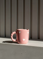Pink mug with heart designs on a windowsill against a gray wall.