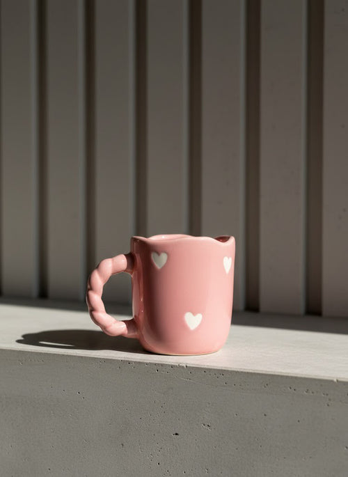 Pink mug with heart designs on a windowsill against a gray wall.
