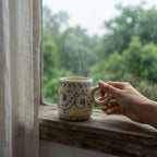 A ceramic mug with a hand-painted owl design sits on a rustic wooden windowsill, with a person's hand holding its handle. Steam rises from the mug, and a blurred green outdoor garden is visible through the rain-speckled window behind it.