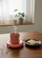Pink cup on a matching pink coaster with steam, next to a plate of cookies on a wooden table.