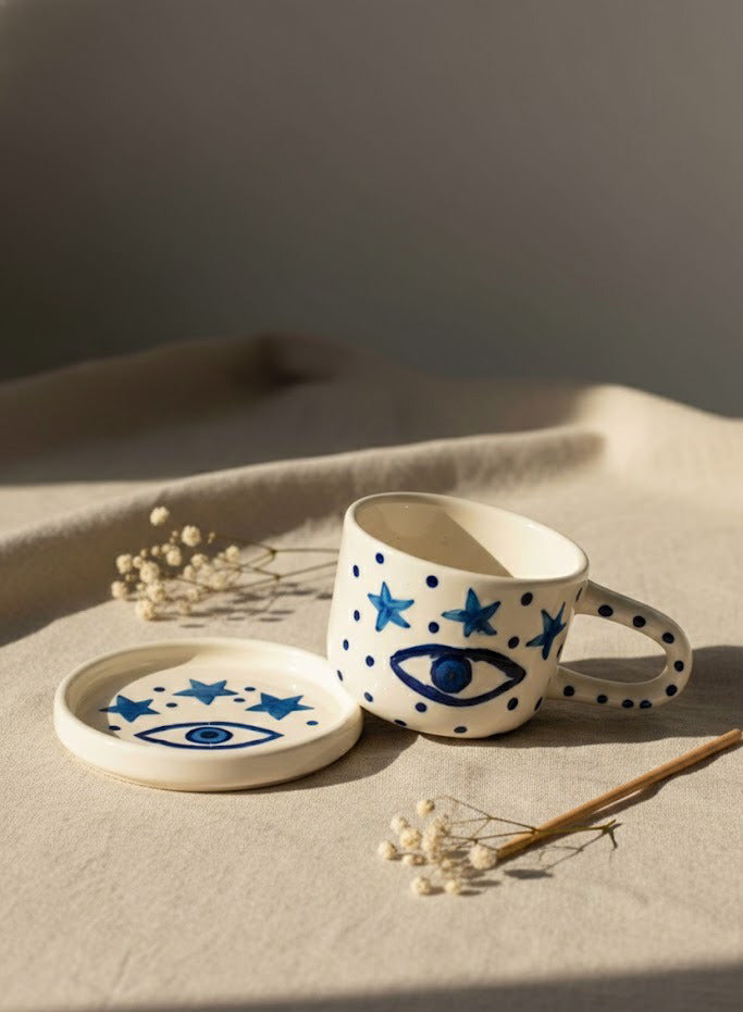 Ceramic mug and coaster with star and eye designs on a neutral background