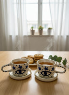 Two ceramic cups with star and eye design on saucers, placed on a wooden table with cookies in the background.