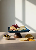 Blue platter with cheese, fruits, and bread on a wooden stand against a white wall.