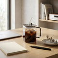Glass coffee mug with iced coffee on a wooden desk with a laptop, notebook, and pen.