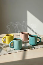 Four ceramic mugs in different colors (yellow, pink, and two greens) on a light surface with steam rising from them.