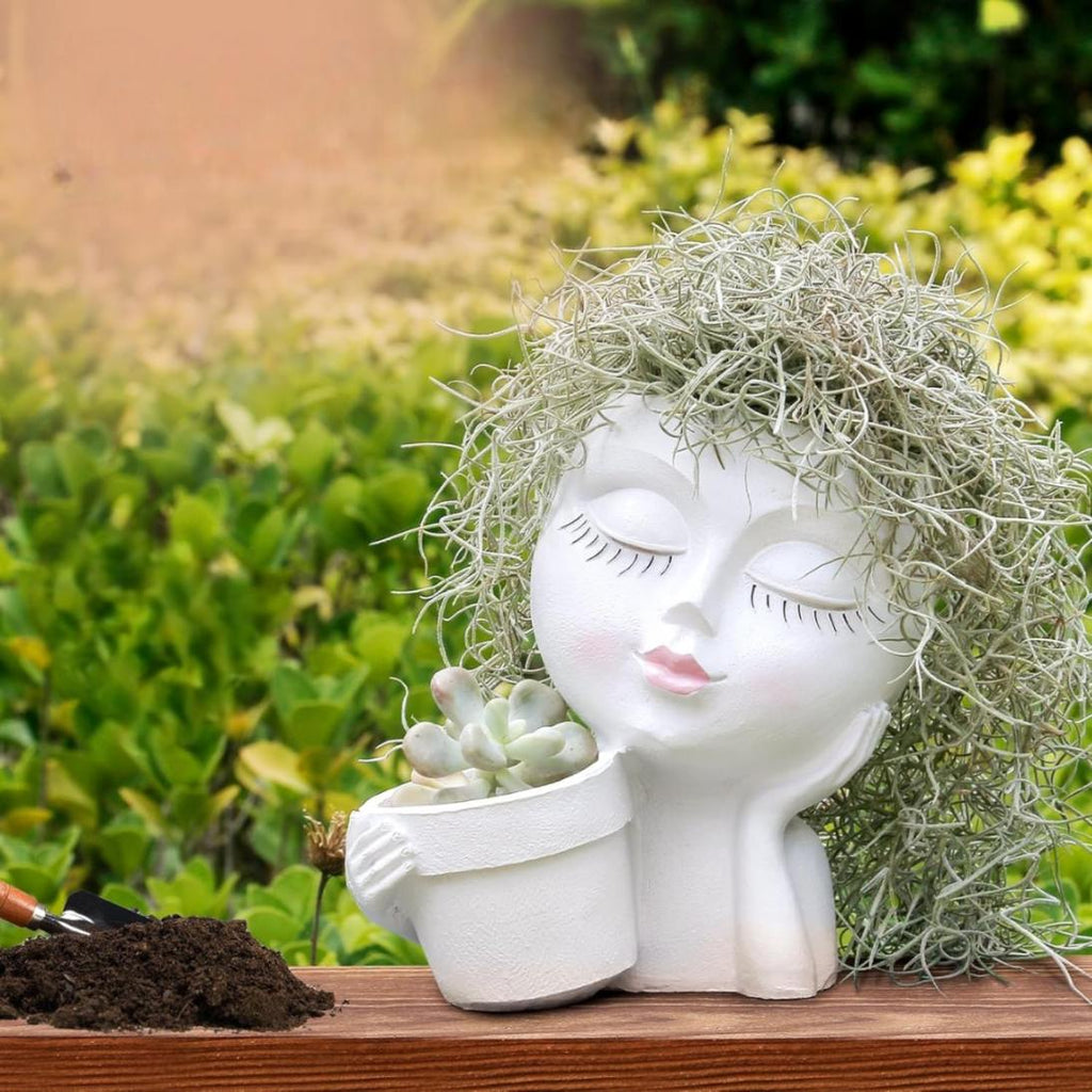 Decorative planter shaped like a face with closed eyes and a pot of succulents, set against a garden backdrop.