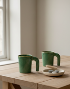 Two green ceramic mugs on a wooden table with a small dish of stones.