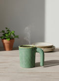 Green ceramic mug with steam on a wooden table, potted plant and plates in the background