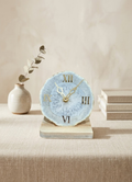Decorative clock with Roman numeral design on a wooden base, placed on a surface with a neutral background.