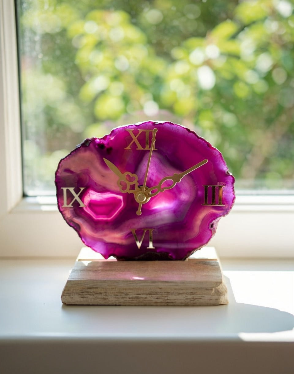 Pink agate clock with gold Roman numeral hands on a wooden base, placed by a window.