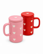 Two ceramic mugs with lids, one pink and one red, both decorated with small white heart cutouts, shown on a white background.