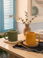 Two mugs on a table with a vase and small plant in the background