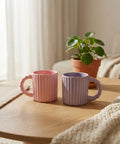 Two ceramic mugs, one pink and one purple, on a wooden surface with a plant in the background.