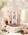 A glass jar candle labeled 'evasa Iced Latte Candle' filled with light beige wax and dark brown coffee beans, styled on a stone slab with coffee beans, vanilla bean, caramel, ice cubes, and a small glass pitcher of milk in the background.