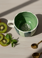 Green ceramic mug with a kiwi design on a light wooden surface with kiwis and a spoon.