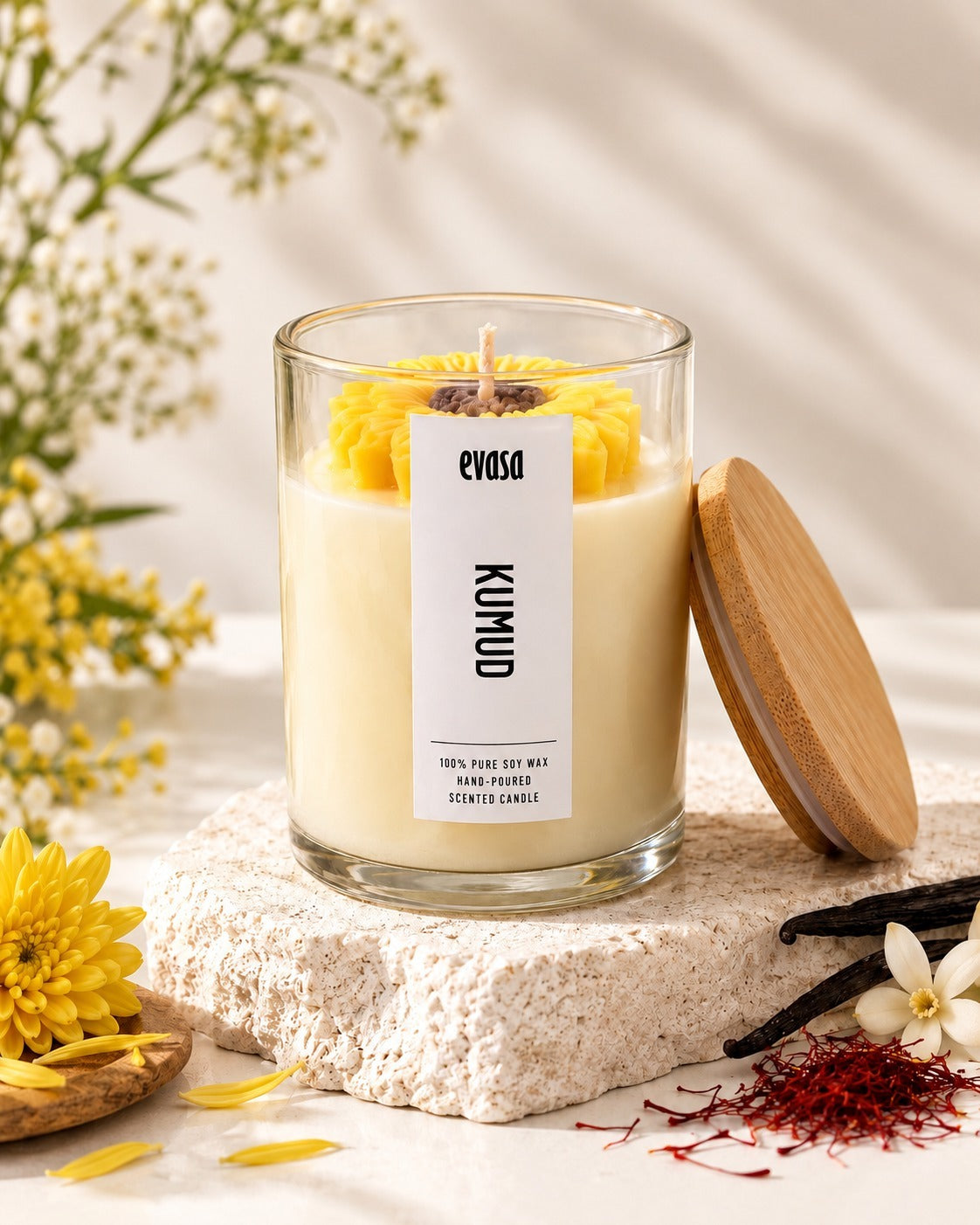 A glass jar candle with creamy off-white wax and a yellow flower on top, labeled 'evasa KUMUD', styled on a stone slab with a wooden lid leaning against it, surrounded by yellow flowers, vanilla beans, and saffron threads.
