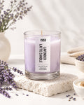 A lavender latte candle in a clear glass jar sits on a textured stone slab, surrounded by sprigs of lavender, a small bowl of honey, and soft neutral props in a bright, clean setting. The candle wax is light lavender purple with a white label reading 'eyasa' and 'LAVENDER LATTE CANDLE'.