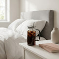 Iced coffee in a glass mug with a straw on a nightstand next to a bed.