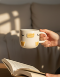Person holding a mug with a face design while reading a book indoors.