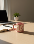 Pink 'LOVE' mug on a desk with a laptop, notebook, and plant in the background.