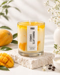 A glass jar candle labeled 'MANGO LATTE CANDLE' by evas, styled on a textured stone slab with fresh mangoes, green leaves, white flowers, and coffee beans in a bright, clean setting.