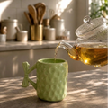 Green textured mug being filled with tea from a clear teapot on a kitchen counter.