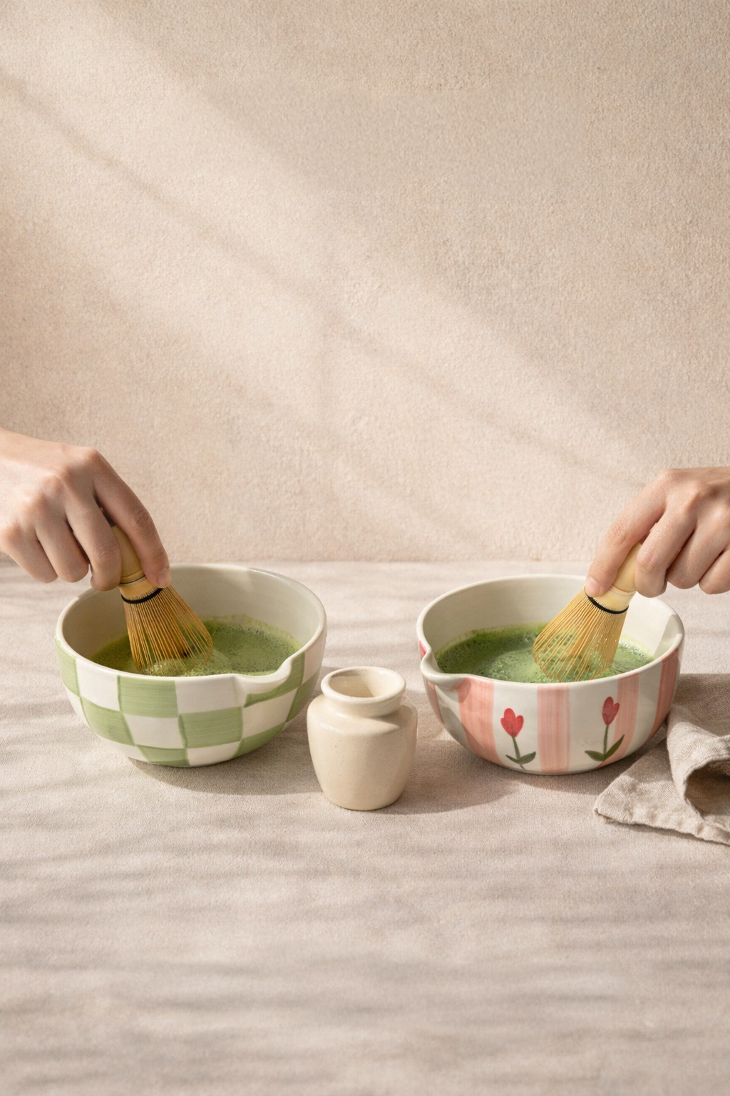 Two hands using wooden whiskes to mix green tea in ceramic bowls on a textured surface.