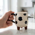 Hand holding a cow-patterned mug on a marble surface with a blurred kitchen background