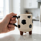 Hand holding a cow-patterned mug on a marble surface with a blurred kitchen background