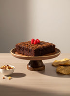Chocolate cake on a wooden stand with raspberries, surrounded by cookies and a bowl of nuts on a wooden surface.