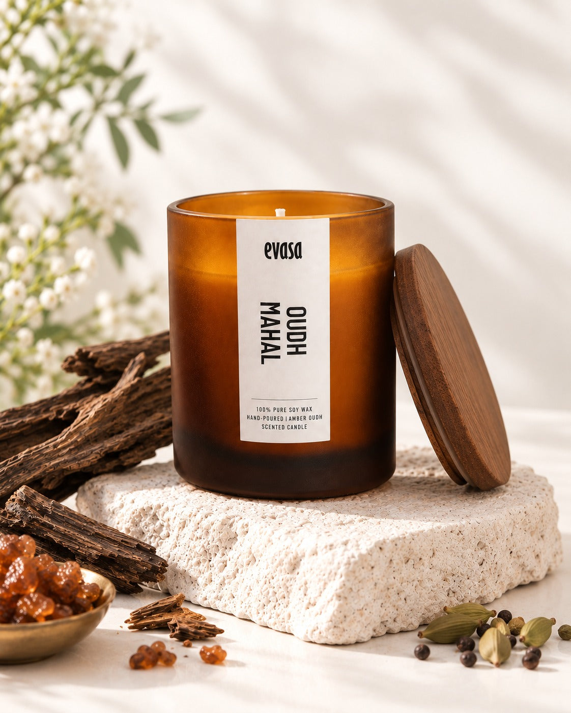 An amber glass candle jar labeled 'evasa OUDh MAHAL' sits on a textured white stone block, with its wooden lid leaning against it. The scene is styled with warm spices, dried bark, and soft white flowers in the background.