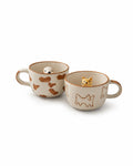 Two ceramic cat-themed mugs on a white background. One mug features brown cat silhouettes and a small white cat figurine inside, while the other features brown outline cat drawings and a small orange tabby cat figurine inside.