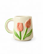 An off-white ceramic mug featuring a raised relief design of two pink tulips with green stems and leaves, and a green handle.