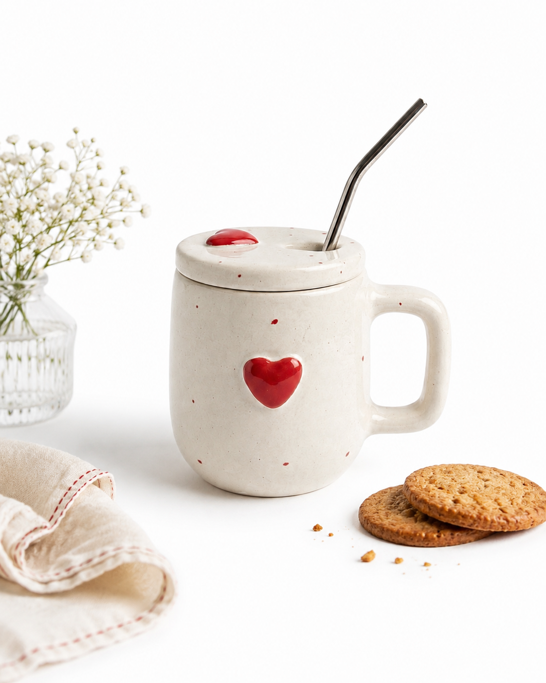 A speckled off-white ceramic mug with a matching lid, a red heart detail, and a metal straw, styled on a white surface with cookies, a napkin, and baby's breath flowers.