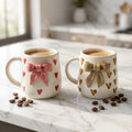 Two ceramic mugs with decorative bows and heart patterns on a marble surface.