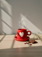Red mug with a heart design on a saucer, surrounded by cookies and beans on a wooden surface with sunlight casting shadows.