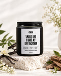 A black scented candle jar with a black lid and a white label reading 'eyasa SMELLS LIKE I HAVE MY LIFE TOGETHER' and 'VANILLA + PATCHOULI + MUSK', styled on a stone slab with vanilla beans, white flowers, and cinnamon sticks.