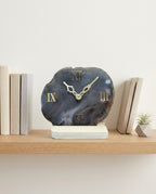 Decorative clock with Roman numeral design on a shelf with books and a plant.