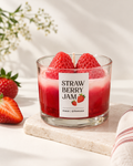 A glass jar candle labeled 'STRAWBERRY JAM' filled with red wax and topped with two whole strawberries, styled on a light stone slab with fresh strawberries and a striped cloth nearby.