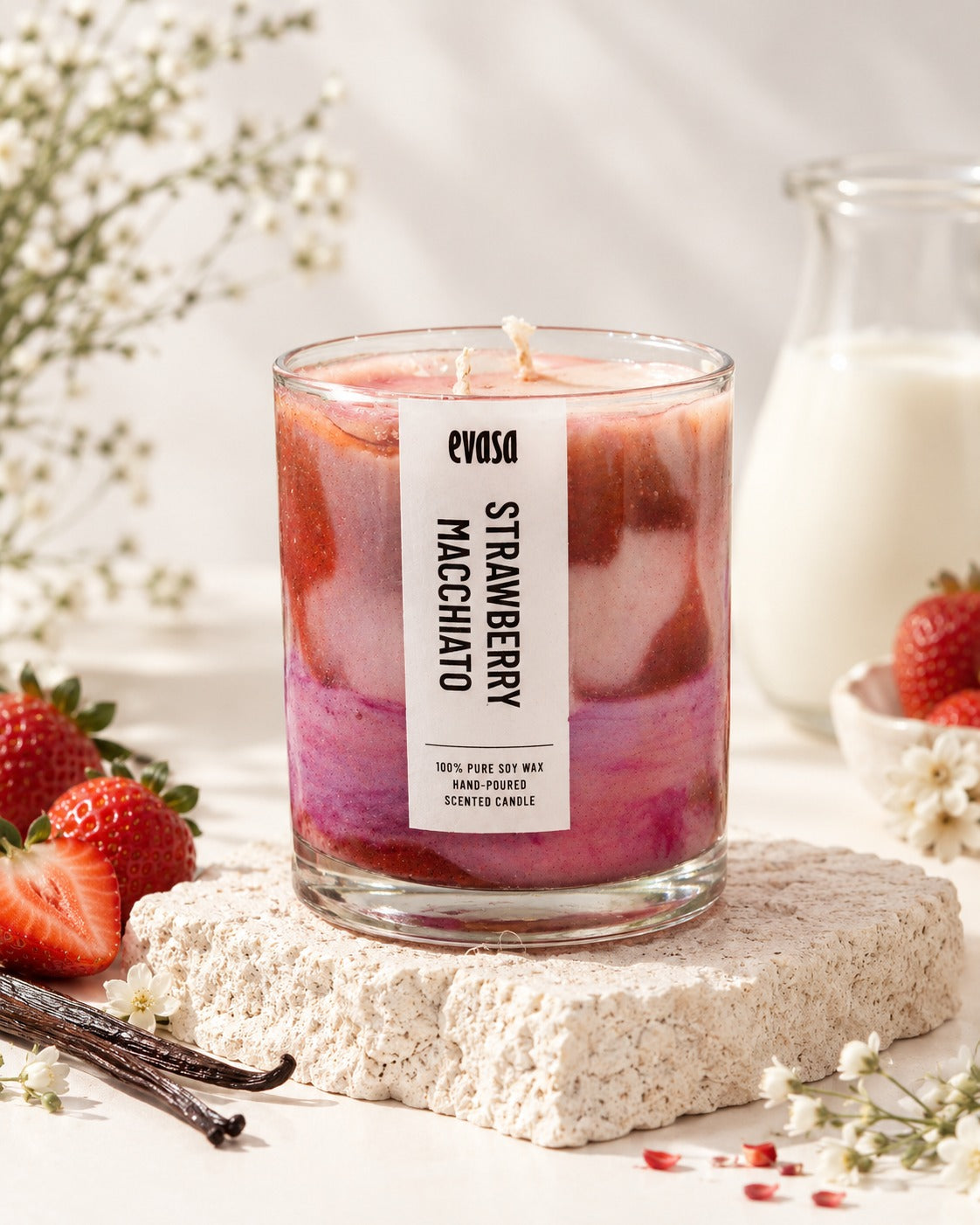 A strawberry macchiatto scented candle by evva's in a clear glass jar, styled on a textured stone block with fresh strawberries, vanilla beans, and a glass of milk in the background.