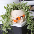 Decorative planter with a smiling face playing a guitar, surrounded by green leaves.