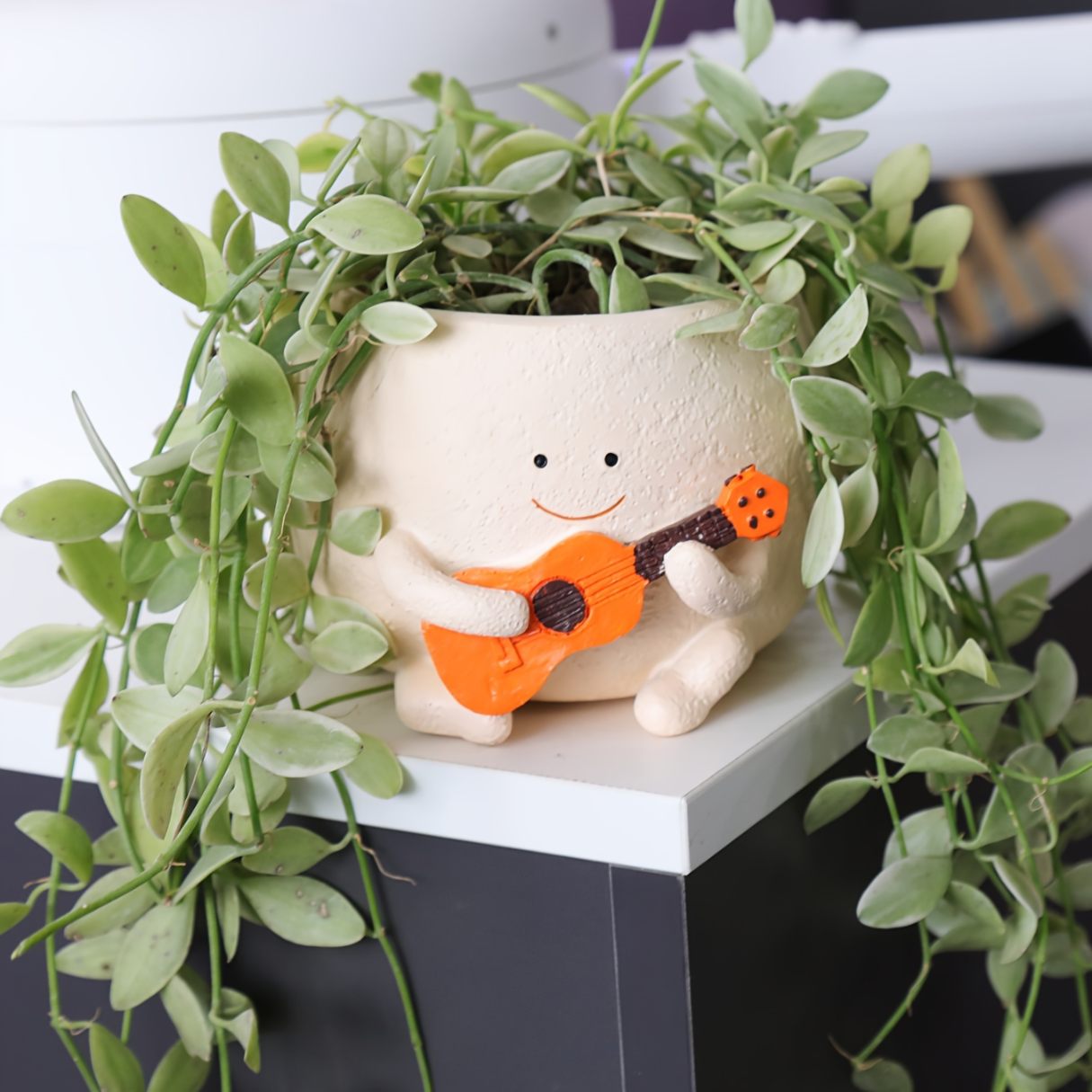 Decorative planter with a smiling face playing a guitar, surrounded by green leaves.