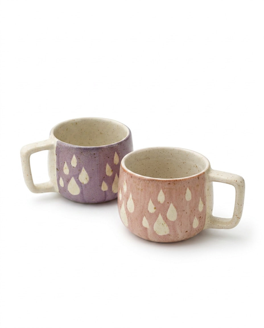Two small ceramic mugs with rounded handles, one in lavender purple and one in dusty pink, both decorated with cream-colored teardrop motifs on a white background.