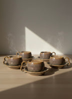 Set of six brown ceramic cups and saucers on a wooden surface with a soft shadow.
